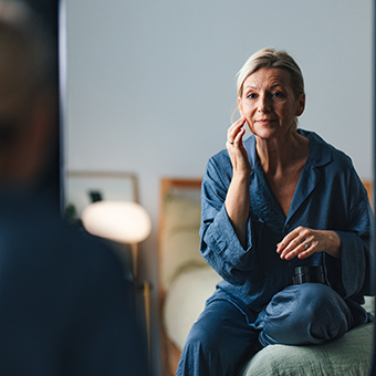 older woman sitting on the bed in her bedroom and looking at her reflection.