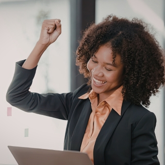 Woman in business attire looking a laptop screen smiles and holds her hand up in a cdelebratory gesture