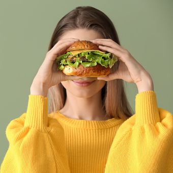 A woman wearing a yellow shirt holding a chicken sandwich to her face.